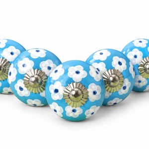May include: Five round cabinet knobs with a bright blue base color and a hand-painted white flower pattern featuring dark blue centers. Each knob has a polished silver-toned metal hardware fitting at the center with a fluted, circular base.
