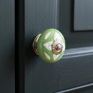 May include: A round ceramic cabinet knob featuring a sage green base with a white leaf pattern. The centre of the knob is accented with a scalloped, metallic silver-toned metal plate and a small, raised circular silver-toned centre stud.