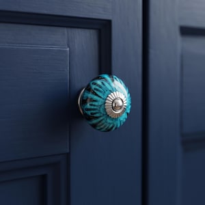 May include: A round ceramic cabinet knob featuring a vibrant turquoise and black floral pattern. The center of the knob is accented with a polished silver-tone metal cap with a fluted edge, mounted on a dark navy blue cabinet door.