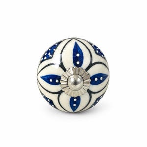 May include: A round ceramic cabinet knob featuring a cream-colored base with a hand-painted blue floral pattern, black outlines, and white dot accents. The center of the knob is adorned with a metallic silver-toned fluted circular metal cap.