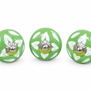 May include: Three round ceramic cabinet knobs featuring a glossy light green finish with hand-painted white leaf patterns. Each knob is centered with a polished silver-tone metal hardware cap that has a scalloped edge and a smooth rounded top.