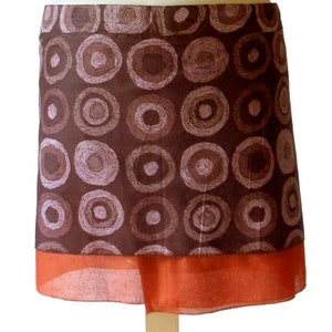 Può includere: Una minigonna marrone con un bordo in tessuto arancione e rosa a strati. La gonna ha un motivo di cerchi bianchi con un centro marrone.