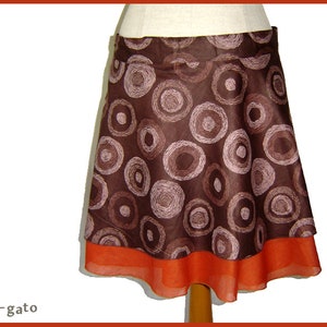 Può includere: Una gonna marrone con un motivo a cerchi rosa e bianchi. La gonna ha uno strato superiore marrone e uno strato inferiore arancione.