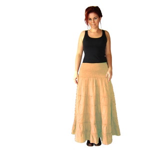 May include: A long, tiered skirt in a light beige colour. The skirt has a smocked waistband and is made of a soft, flowing fabric.