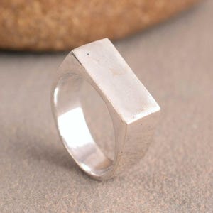 Pode incluir: Um anel de metal prateado com uma superfície superior plana e retangular. A banda é sólida e estreita ligeiramente em direção à base, apresentando um acabamento polido com reflexos subtis em toda a superfície exterior do anel.