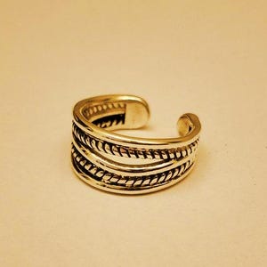 Puede incluir: Un anillo abierto de color plateado con un diseño de múltiples bandas en tres filas distintas. Las filas exteriores muestran una textura de cuerda trenzada, mientras que la banda central es lisa. El anillo tiene una abertura ajustable en la parte posterior.