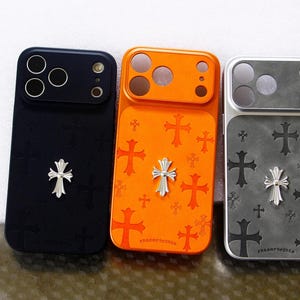 Puede incluir: Tres fundas rectangulares para teléfono en negro, naranja y gris. Cada funda presenta un patrón de cruces repetidas y un emblema de cruz plateado prominente centrado en la parte posterior. El área de la cámara tiene aberturas circulares.