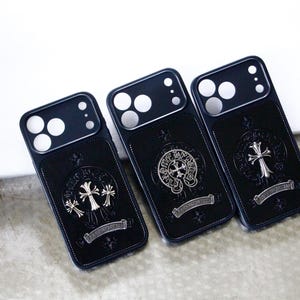 May include: Three black smartphone cases featuring a rectangular design with a camera cutout at the top. Each case displays a central silver-toned metallic emblem shaped like a cross, surrounded by circular decorative motifs and a small banner.