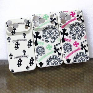 Puede incluir: Tres fundas blancas para smartphone con motivos de cruces en negro, verde y rosa, y emblemas circulares con el texto Chrome Hearts. Cada funda tiene un recorte rectangular para la cámara y un patrón repetitivo de símbolos gráficos.