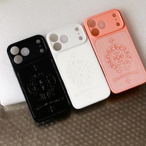 May include: Three smartphone cases in black, white, and peach, each featuring a circular embossed logo with the text CHROME HEARTS. The cases have cutouts for a triple camera system and flash, with a rectangular design and a cross symbol at the top.