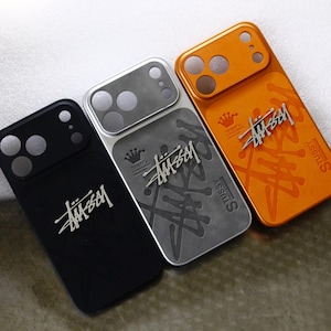Puede incluir: Tres fundas para smartphone en negro, plata y naranja, cada una con un área de recorte para la cámara, un icono de corona y el logotipo de Stussy en una fuente cursiva estilizada. Las fundas tienen un acabado liso con patrones sutiles.