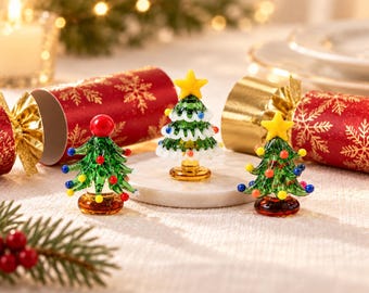 May include: Three miniature glass Christmas tree figurines. The centre tree features a yellow star topper and white garland. The two flanking trees are adorned with colourful glass spheres, with one topped by a red sphere and the other by a yellow star.