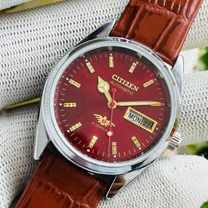 Può includere: Un orologio da polso automatico Citizen con cassa rotonda color argento, quadrante rosso intenso con indici dorati e lancette dorate. Il quadrante riporta la scritta CITIZEN AUTOMATIC, 21 JEWELS e una finestrella giorno-data. Cinturino in pelle marrone.