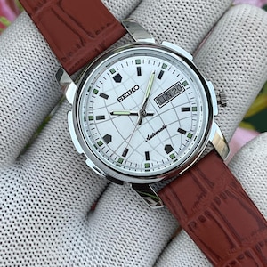 Peut inclure: Une montre automatique Seiko avec un boîtier rond argenté et un cadran blanc à motif quadrillé. La montre possède des aiguilles argentées, des index verts, un guichet jour-date à trois heures et un bracelet en cuir marron.