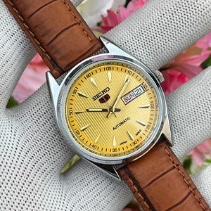 Pode incluir: Um relógio de pulso automático Seiko 5 com caixa redonda de aço inoxidável prateado e mostrador dourado texturizado. O mostrador apresenta ponteiros e índices dourados e uma janela de dia e data na posição das três horas. Pulseira de couro castanho.