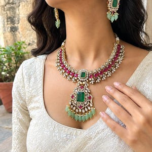 May include: A gold-toned necklace and matching drop earrings set featuring rectangular green center stones, vibrant pink oval accents, and small white pearls. The necklace includes a large central pendant with dangling light green beads.