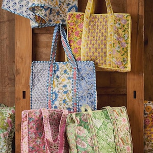 May include: A collection of four quilted tote bags featuring floral block print patterns in yellow, blue, pink, and green. Each bag has two shoulder straps and a rectangular shape with piped edges. A matching quilted fabric piece hangs above the bags.