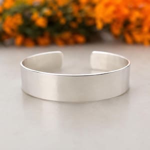 May include: A wide, polished silver cuff bracelet with a smooth, reflective surface and a clean, open-ended design. The bracelet features a simple, unadorned band that curves into a circular shape, showcasing a bright, metallic finish throughout.
