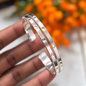 Puede incluir: Una pulsera de brazalete de plata pulida con un diseño de doble banda. La superficie del metal está acentuada con una serie de hendiduras circulares espaciadas uniformemente que crean una apariencia texturizada a lo largo de las bandas.