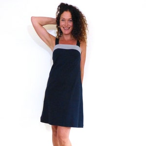 Puede incluir: Una mujer con un vestido negro sin mangas con una banda gris en el pecho. Está de pie sobre un fondo blanco y sonriendo.