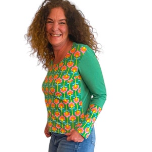 Könnte beinhalten: Eine Frau trägt ein grünes Langarmshirt mit einem Blumenmuster auf der Vorderseite und den Ärmeln. Das Shirt hat einen runden Ausschnitt und eine figurbetonte Silhouette.