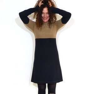 Puede incluir: Un vestido de bloques de color con una sección superior color canela y una sección inferior negra. El vestido tiene mangas largas con coderas negras. La modelo lleva medias negras y botas grises con cordones.