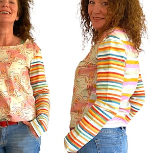 May include: A white long-sleeved shirt with a colorful striped pattern on the sleeves. The shirt has a pink and white pattern on the front with a repeated image of a pink bird with the word "Duckling" written on it.