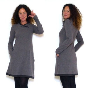 May include: A gray long-sleeved dress with a cowl neck and a contrasting black trim. The dress is shown on a model wearing black tights and black boots.