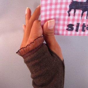 Puede incluir: Un guante sin dedos de punto marrón con un borde de encaje marrón. El guante está siendo sostenido por una mano que sostiene una tela de cuadros rosa y blanca con una silueta negra de un reno y la palabra "Sit" impresa en ella.