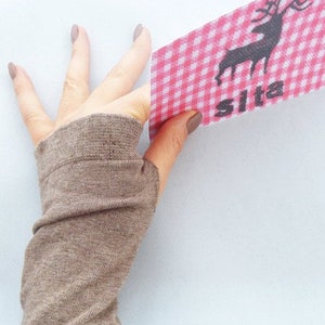 Puede incluir: Un calientamanos marrón sin dedos hecho de tejido de punto. El calientamanos se muestra en una mano que sostiene una tela de cuadros rojos y blancos con una silueta negra de un ciervo y la palabra "Sita" impresa en ella.