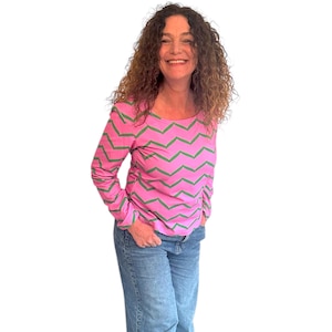 May include: A woman wearing a pink and green striped long-sleeve shirt with a chevron pattern. She is wearing blue jeans and has curly brown hair.