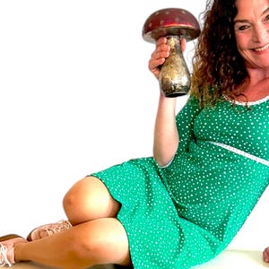 Puede incluir: Una mujer con un vestido verde con lunares blancos se sienta en una superficie blanca. Está sosteniendo un jarrón grande en forma de hongo rojo y dorado.