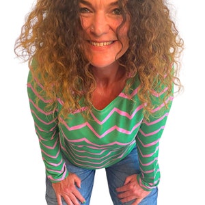 May include: A woman wearing a green and pink striped long-sleeved shirt with a chevron pattern. She is wearing blue jeans and is kneeling with her hands on her knees.