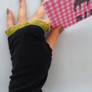 Puede incluir: Guantes negros sin dedos con un borde de encaje verde. Los guantes se llevan en una mano con esmalte de uñas marrón.