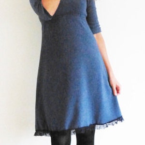 May include: A woman wearing a blue dress with a lace trim and black tights. She is standing in front of a white wall.