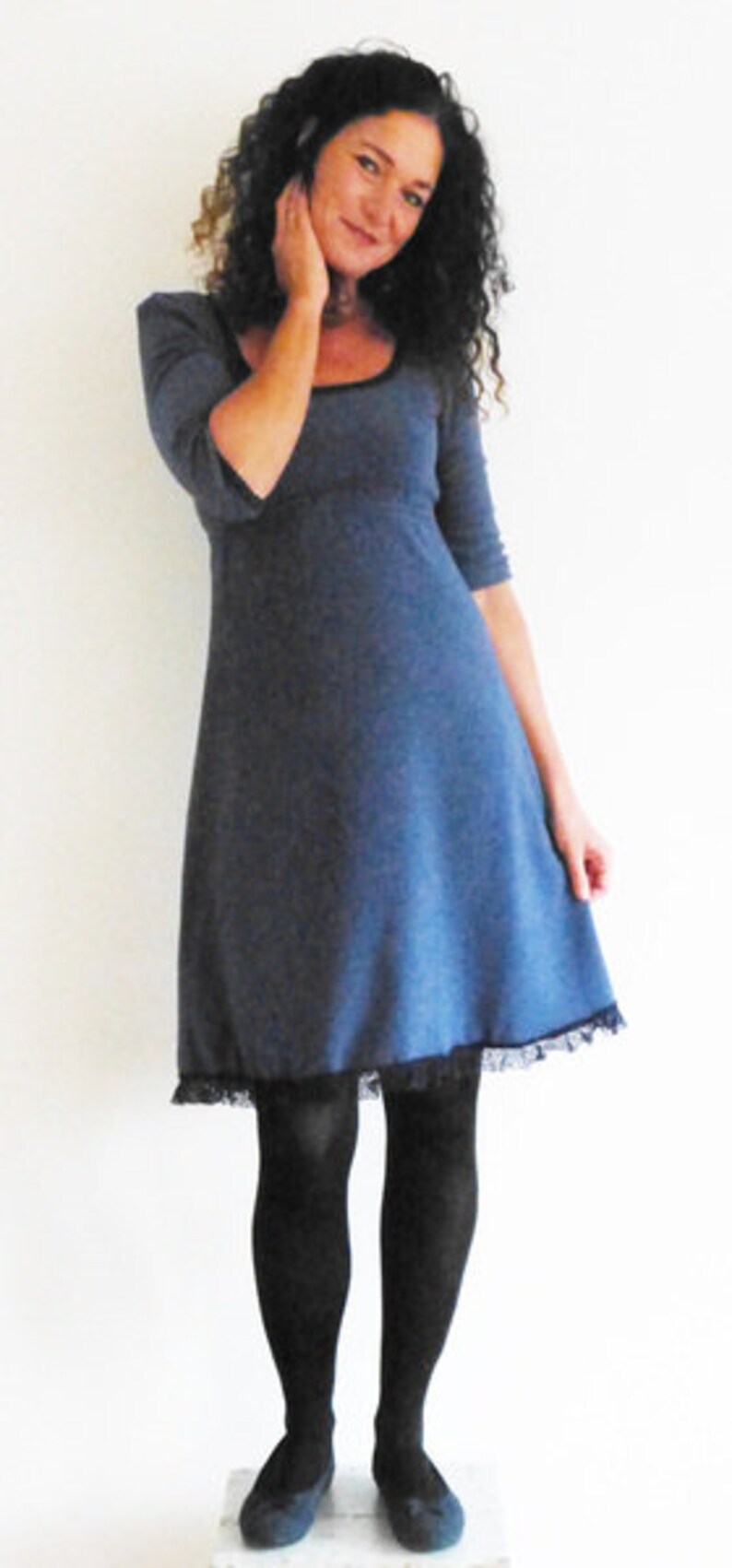 May include: A woman wearing a blue dress with a lace trim and black tights. She is standing in front of a white wall.