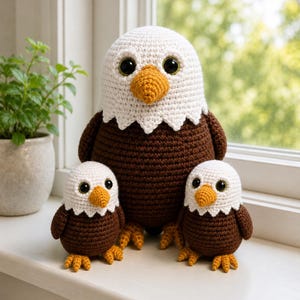 May include: Three crocheted bald eagle figures featuring white heads, dark brown bodies, and bright yellow beaks and feet. The largest eagle stands in the center, flanked by two smaller, identical eagle figures, all with large black safety eyes.