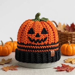 May include: A chunky knit beanie designed to look like a jack-o'-lantern, featuring a bright orange pumpkin body with a black stitched face, a green stem on top, and a wide black ribbed brim with a small brown tag labeled NANO KNITS.