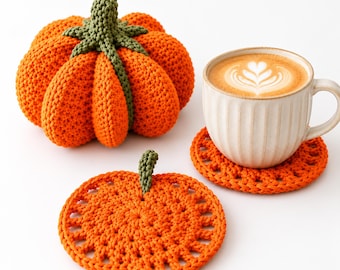Puede incluir: Una calabaza naranja de ganchillo con tallo verde, un posavasos plano con forma de calabaza naranja y una taza de cerámica beige llena de café con arte latte. La taza descansa sobre un segundo posavasos con forma de calabaza, todo con textura.