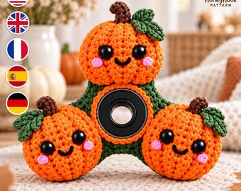 May include: A crochet fidget spinner featuring three orange pumpkins with dark green stems and leaves. Each pumpkin has black plastic eyes, a stitched black mouth, and pink cheeks. A black circular bearing is centered in the green base.