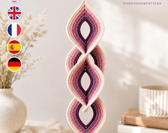 May include: A hanging crochet spiral ornament featuring a gradient color scheme that transitions from cream to light pink, rose, and deep purple. The structure consists of four interconnected leaf-shaped loops, suspended by a single yarn cord.