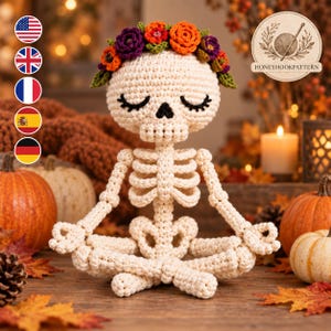 Könnte beinhalten: Eine gehäkelte cremeweiße Skelettfigur in sitzender Meditationspose mit geschlossenen Augen, schwarzen Wimpern und einem Kranz aus orangefarbenen, violetten und grünen gehäkelten Blumen. Der Text HONEYHOOKPATTERN ist oben rechts zu sehen.