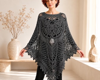 May include: A dark gray crochet poncho featuring an intricate butterfly motif at the center, a geometric open-knit pattern throughout, and a pointed hemline with decorative fringe detailing. The garment is worn over a black top and black pants.