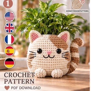 May include: A crochet pattern PDF download featuring a cat-shaped plant pot cover. The cat is cream and brown with black eyes, a pink nose, and a striped tail. A green plant sits inside the crochet cover. Text includes HONEYHOOKPATTERN.
