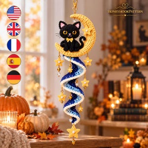 Op de afbeelding: Een gehaakte wandhanger met een zwarte kat die op een gele halve maan zit. Onder de maan hangt een blauw-witte spiraalvormige staart, versierd met kleine gele sterretjes, doorzichtige kralen en gouden accenten. De tekst HONEYHOOKPATTERN is zichtbaar.