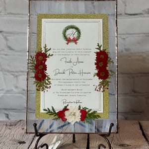 May include: A framed wedding invitation with gold glitter accents and floral embellishments. The invitation features the names "Faith Anne" and "David Peter Heeren" with event details. The frame is displayed on a decorative metal stand.