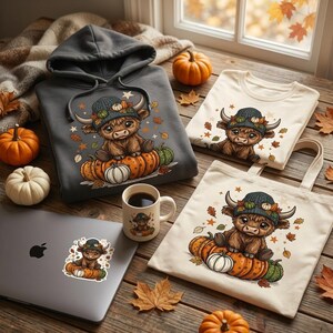 May include: A dark grey hoodie, a cream t-shirt, a cream tote bag, and a white ceramic mug, each featuring a graphic of a brown highland cow wearing a knit hat and sitting on pumpkins, alongside a laptop with a matching sticker.