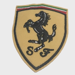 Op de afbeelding: Een schildvormige geborduurde patch met een gele achtergrond en een zwarte silhouet van een steigerend paard in het midden. De letters S en F zijn in het zwart onder het paard geborduurd. Een horizontale groen-wit-rode streep siert de bovenrand.