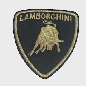 Könnte beinhalten: Ein schildförmiger, bestickter Aufnäher mit schwarzem Hintergrund und einem goldenen Stier-Emblem in der Mitte. Das Wort LAMBORGHINI ist in goldenen Großbuchstaben im oberen Bereich des Aufnähers eingestickt und von einem goldenen Rand umgeben.