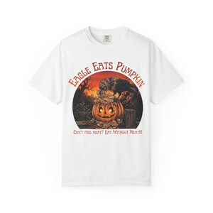 Puede incluir: Una camiseta blanca de manga corta con el gráfico de un pájaro negro posado sobre una calabaza tallada. El texto EAGLE EATS PUMPKIN y CAN'T FIND MEAT? EAT WITHOUT MEAT!!! aparece sobre un fondo de atardecer con árboles y una valla.